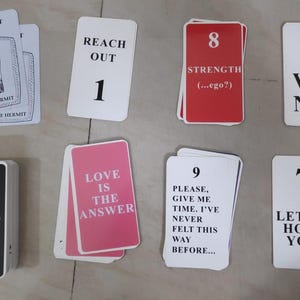 Op de afbeelding: Een verzameling tarotkaarten met verschillende berichten en nummers. Kaarten zijn onder meer "The Hermit", "Reach Out 1", "Strength 8", "Wand 1", "I Want To Get You Drunk 15", "Love Is The Answer", "Please, Give Me Time 9" en "Let Me Hold You 7."
