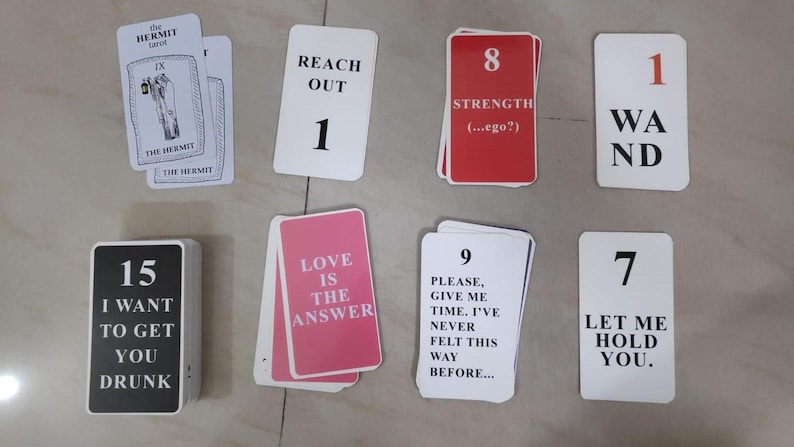 Op de afbeelding: Een verzameling tarotkaarten met verschillende berichten en nummers. Kaarten zijn onder meer "The Hermit", "Reach Out 1", "Strength 8", "Wand 1", "I Want To Get You Drunk 15", "Love Is The Answer", "Please, Give Me Time 9" en "Let Me Hold You 7."