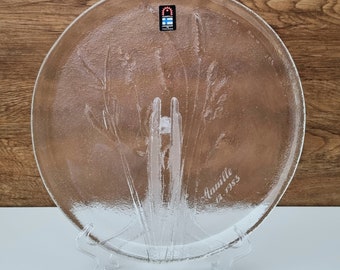 May include: A clear glass serving plate with a raised design of tall grass. The plate is inscribed with the text "Aunuille 12. 1983".