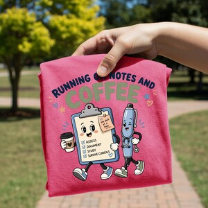 Könnte beinhalten: Ein pinkfarbenes T-Shirt mit einer Grafik eines lächelnden Klemmbretts und eines Stifts mit Stethoskop. Der Text lautet "RUNNING ON NOTES AND COFFEE" und "one shift at a time" auf einem Haftnotizzettel. Das Klemmbrett listet "ASSESS", "DOCUMENT", "STUDY", "SURVIVE CLINICALS".