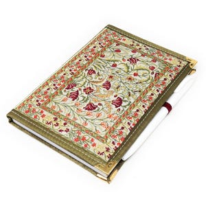 Op de afbeelding: Een decoratief notitieboek met een bloemenpatroon op de omslag. De omslag heeft een crèmekleurige achtergrond met rode en groene bloemmotieven. Het notitieboek heeft een groene rand en gouden hoekbeschermers. Een witte pen met een bordeauxrode band is erin gestoken.