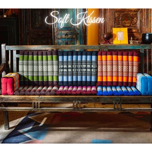 May include: A weathered wooden bench with colorful, patterned cushions. The cushions are in shades of red, green, blue, and orange. The text "Soft Kissen" is displayed above the bench in a neon-style font.