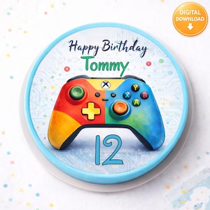 Può includere: Una torta rotonda con un bordo blu con un colorato design a controller di videogiochi. La torta ha la scritta "Happy Birthday Tommy" e il numero "12" in blu. Un'icona di download digitale si trova in alto a destra.