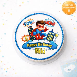 May include: A round birthday cake with blue frosting trim. The cake features a superhero theme with a cartoon illustration of a superhero, the words "Happy Birthday Niki", and the number 5. The words "Pow!" and "Wow!" are also displayed.