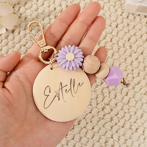 Puede incluir: Un llavero de madera con el nombre "Estelle" grabado en un dije redondo de madera. El llavero presenta un cierre dorado, una flor morada, cuentas de madera y una cuenta geométrica morada.