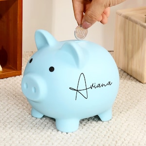 Può includere: Un salvadanaio azzurro con il nome "Ariana" in corsivo nero. Una mano sta inserendo una moneta nella fessura. Il salvadanaio ha occhi neri e quattro piccole zampe. Una piccola statuina di scoiattolo è visibile sullo sfondo.