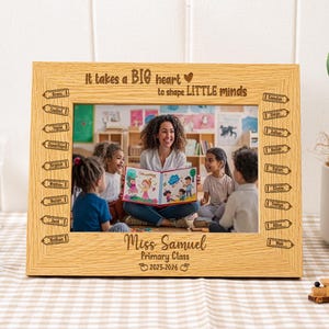 May include: A light oak picture frame with a photograph of a teacher reading to children. The frame is inscribed with the words "It takes a BIG heart to shape LITTLE minds" and "Miss Samuel Primary Class 2025-2026". Pencil illustrations are etched.