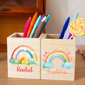 Puede incluir: Dos portalápices de madera con diseños de arcoíris. Uno está etiquetado como "Rachel" y el otro como "Eudora". Los portalápices contienen bolígrafos, lápices y una piruleta. Los portalápices son de madera clara.