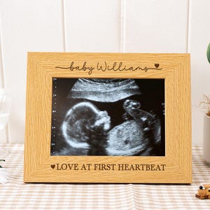 Op de afbeelding: Een licht houten fotolijst met een echografie. De lijst heeft de tekst "baby Williams" en "LOVE AT FIRST HEARTBEAT" gegraveerd. Een kleine cactus en speelgoedtrein staan op de achtergrond.