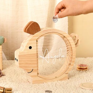 May include: Wooden piggy bank shaped like a pig with a clear front panel. The name "Angelina" is written on the front. A hand is dropping a coin into the slot. Other wooden toys are in the background.