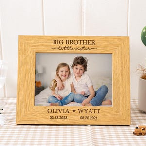 May include: A light oak picture frame with the words "BIG BROTHER little sister" engraved at the top. The frame holds a photo of two children. The names Olivia and Wyatt are below the photo, along with their birthdates.