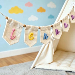 May include: A decorative banner featuring six off-white canvas pennants strung on a cord with wooden beads. Each pennant displays a single lowercase letter in a different colored yarn, spelling out the name lennon in pink, yellow, and blue.