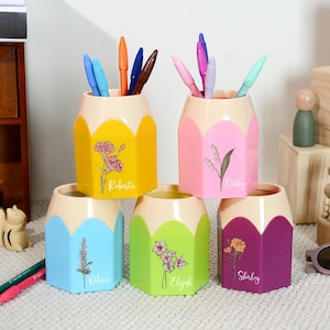 Puede incluir: Cinco portalápices con forma de lápiz en amarillo, rosa, azul, verde y morado. Cada soporte tiene una parte superior color crema y contiene bolígrafos. Diseños florales y nombres están impresos al frente.