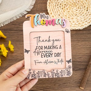 Puede incluir: Un marco de fotos de madera con el texto "Thank you for making a difference every day" y "From Alison White". El marco tiene un borde floral y el nombre "Mrs. Thomas" en letras coloridas.