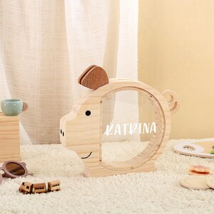 May include: A wooden piggy bank shaped like a pig with a clear front, featuring the name "KATRINA" in white. The bank has a slot on top and a small wooden ear and tail. The design is simple and child-friendly.