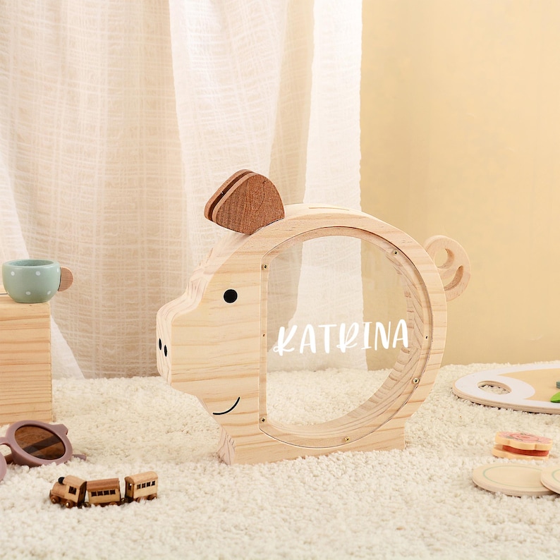 May include: A wooden piggy bank shaped like a pig with a clear front, featuring the name "KATRINA" in white. The bank has a slot on top and a small wooden ear and tail. The design is simple and child-friendly.