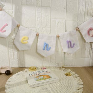 May include: A white fabric banner featuring individual pennants with pastel-colored knitted letters spelling out a name, suspended by wooden beads. Below, a book titled Guess How Much I Love You rests on a round, cream-colored woven mat.