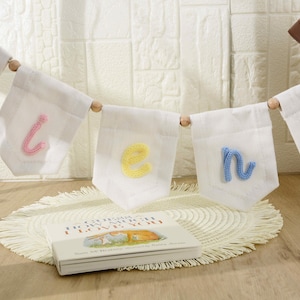 May include: A white fabric banner featuring individual pennants strung together with wooden beads. Each pennant displays a single lowercase letter in a different pastel yarn color, including pink, yellow, and blue, spelling out a name.