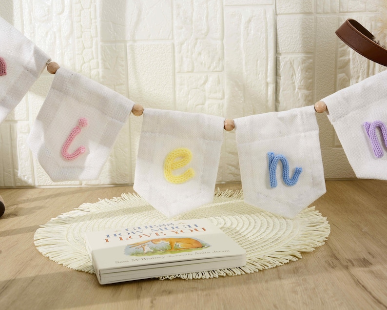 May include: A white fabric banner featuring individual pennants strung together with wooden beads. Each pennant displays a single lowercase letter in a different pastel yarn color, including pink, yellow, and blue, spelling out a name.