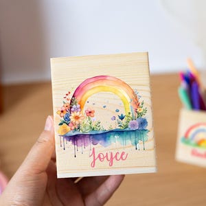 Puede incluir: Una caja de madera con un diseño de arcoíris en acuarela, flores y el nombre "Joyce" en escritura rosa. El arcoíris se extiende por la parte superior de la caja, con gotas coloridas debajo. Una segunda caja con un diseño de arcoíris está en el fondo.