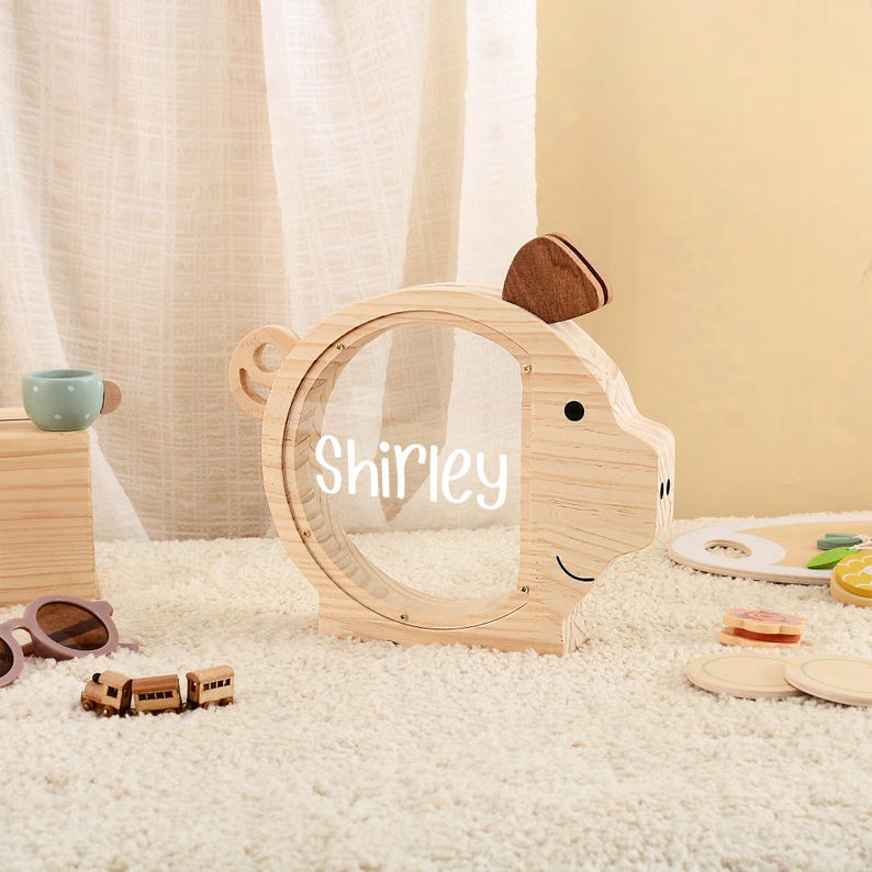 May include: A wooden piggy bank shaped like a pig with a clear center, the name "Shirley" is written on the clear center. The bank has a small wooden ear and a small wooden tail. The bank is sitting on a white rug.