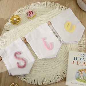 May include: A decorative banner featuring three white fabric pennants strung together with wooden beads. Each pennant displays a single, textured, knitted letter in pink, light pink, and yellow, spelling out the letters S, i, and e.