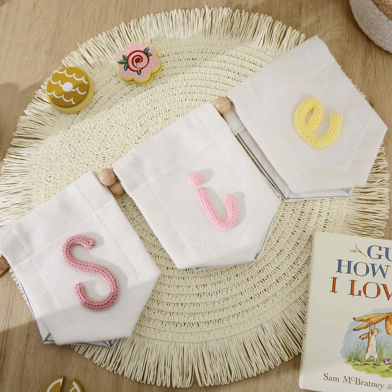 May include: A decorative banner featuring three white fabric pennants strung together with wooden beads. Each pennant displays a single, textured, knitted letter in pink, light pink, and yellow, spelling out the letters S, i, and e.