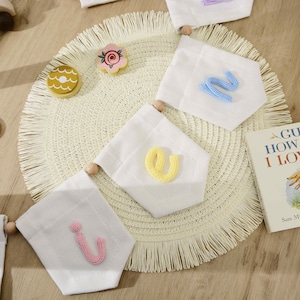 May include: A white fabric banner set featuring three pennants with knitted letters in light blue, yellow, and pink. The pennants are strung together with wooden beads. A small wooden flower and a book titled Guess How Much I Love You are visible.