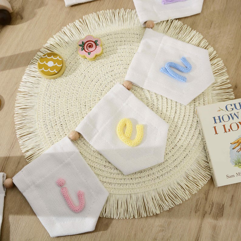 May include: A white fabric banner set featuring three pennants with knitted letters in light blue, yellow, and pink. The pennants are strung together with wooden beads. A small wooden flower and a book titled Guess How Much I Love You are visible.