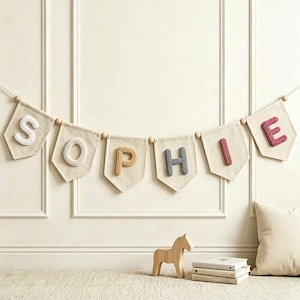 May include: A decorative banner featuring six off-white fabric pennants strung together with wooden beads. Each pennant displays a single, raised, knitted letter in a different color, spelling out the name SOPHIE in white, tan, blue, and pink.
