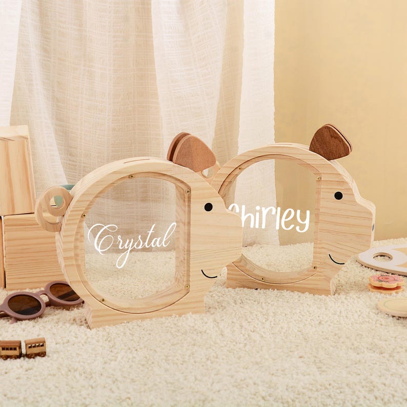 May include: Two wooden piggy banks shaped like pigs, with clear acrylic windows. One bank has the name "Crystal" and the other has "Shirley" written on it. The banks are light brown with brown ear accents and black dot eyes.