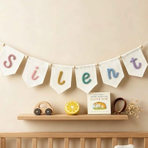 May include: A decorative banner featuring six white fabric pennants suspended on a string with wooden beads. Each pennant displays a single embroidered letter in pink, yellow, or blue, spelling the word silent. The letters are textured.