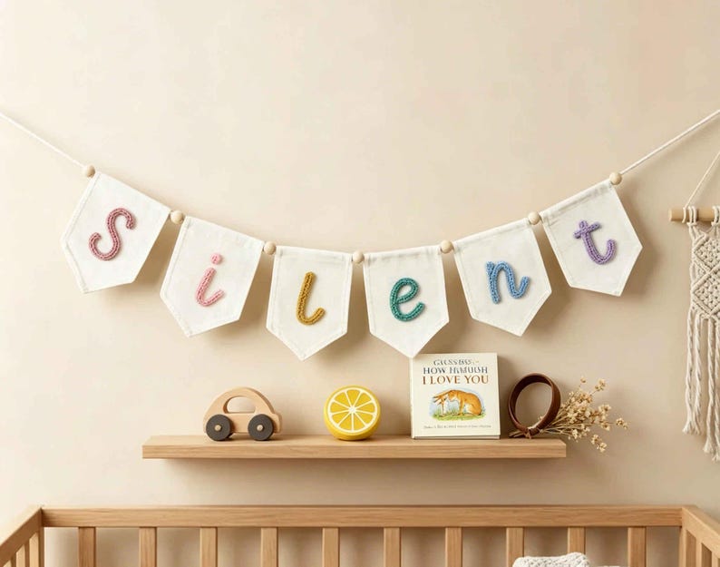 May include: A decorative banner featuring six white fabric pennants suspended on a string with wooden beads. Each pennant displays a single embroidered letter in pink, yellow, or blue, spelling the word silent. The letters are textured.