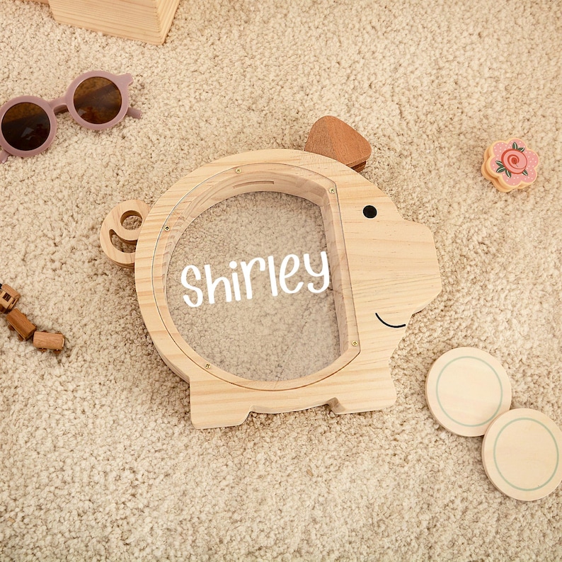 May include: A wooden piggy bank shaped like a pig with a clear front, the name "Shirley" is written in white. The bank has a small tail and ear. Other items include sunglasses and wooden toys.