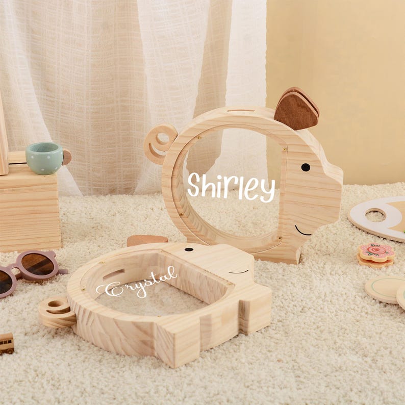 May include: Two wooden piggy banks shaped like pigs, with clear circular windows. One bank has the name "Shirley" and the other "Crystal" written on the window. The banks are light brown with brown ear accents.