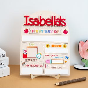 Può includere: Un cartello in legno per il primo giorno di scuola, personalizzato con il nome "Isabella's" in rosso. Il cartello include spazi per scrivere "I am", "Date", "Years Old", "My Teacher Is" e "Things I Love". Elementi decorativi: mele, un arcobaleno e una matita.