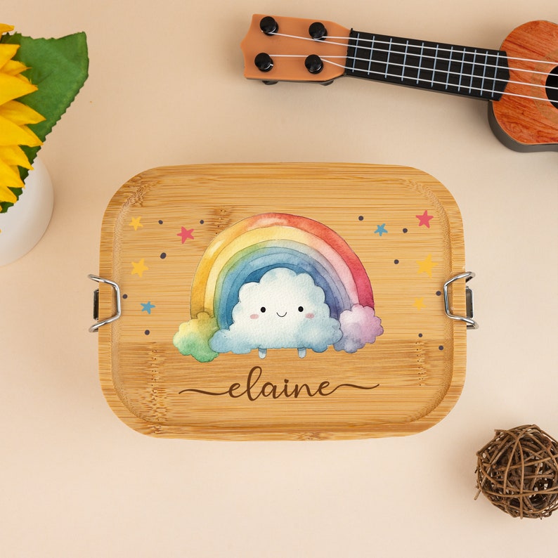 May include: A wooden lunchbox with a rainbow and a cloud with a smiling face. The lunchbox is decorated with stars and the name "elaine" is written on the bottom.