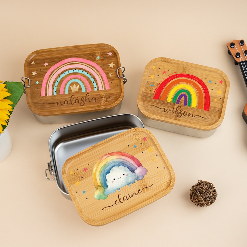 May include: Three personalized bamboo lunchboxes with rainbow designs. The lunchboxes have stainless steel interiors. The first lunchbox has a rainbow with a crown and the name "natasha" written below. The second lunchbox has a rainbow with the name "wilson" written below. The third lunchbox has a rainbow with a cloud and the name "elaine" written below.