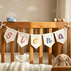 May include: A decorative banner featuring five off-white fabric pennants hanging from a wooden crib. Each pennant displays a single, colorful, crocheted letter spelling the name SIENA in pink, yellow, and blue yarn, secured with white cord.