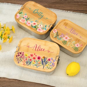 Pode incluir: Três lancheiras personalizadas com tampas de bambu e bases cinzentas. As tampas são decoradas com desenhos florais e nomes: "Betty" em turquesa e "Alice" em rosa. As caixas são retangulares com fechos metálicos.