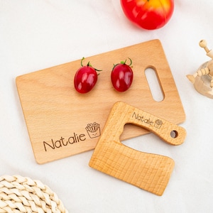 May include: A wooden cutting board and a matching cleaver with the name "Natalie" and a french fry graphic. Two red cherry tomatoes sit on the board. The cutting board has a handle cutout.