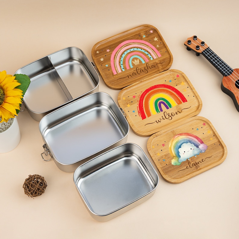 May include: Three stainless steel lunch boxes with a divider in the top box. The lunch boxes are open and stacked on a beige surface. Each lunch box has a matching wooden lid with a rainbow design and a name written on it. The names are Natasha, Wilson, and Elaine.