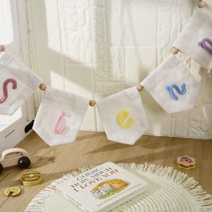 May include: A white fabric bunting banner featuring individual pennants with embroidered letters in pink, yellow, and blue, separated by wooden beads. The letters spell out the word "Sienna" in a soft, textured yarn script style.