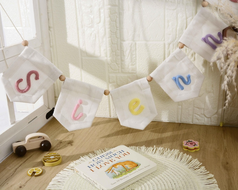 May include: A white fabric bunting banner featuring individual pennants with embroidered letters in pink, yellow, and blue, separated by wooden beads. The letters spell out the word "Sienna" in a soft, textured yarn script style.
