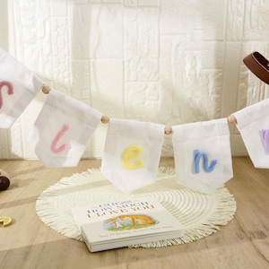 May include: A decorative banner featuring five white fabric pennants strung on a cord with wooden beads. Each pennant displays a single lowercase letter in a different color: pink, light pink, yellow, light blue, and light purple yarn embroidery.
