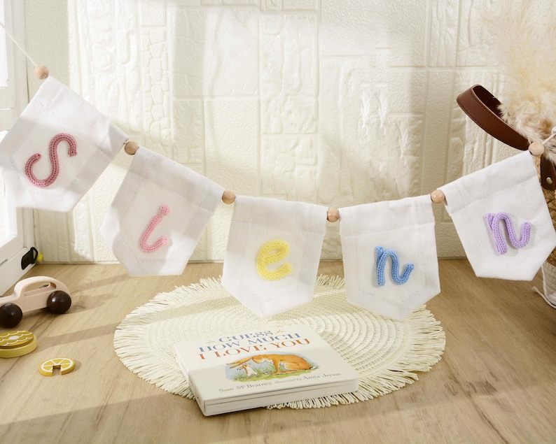 May include: A decorative banner featuring five white fabric pennants strung on a cord with wooden beads. Each pennant displays a single lowercase letter in a different color: pink, light pink, yellow, light blue, and light purple yarn embroidery.