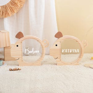 May include: Two wooden piggy banks shaped like pigs, each with a clear circular window. One bank has "Barbara" and the other has "Katrina" written on it. The banks have brown ears and a smiling face.