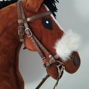 May include: Brown and white plush rocking horse with a black mane and tail. The horse has a brown leather bridle with a silver buckle and a white fur patch on its nose.