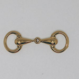 May include: Gold-toned metal horse bit buckle. The buckle has two rings and a centre piece with a rounded shape.