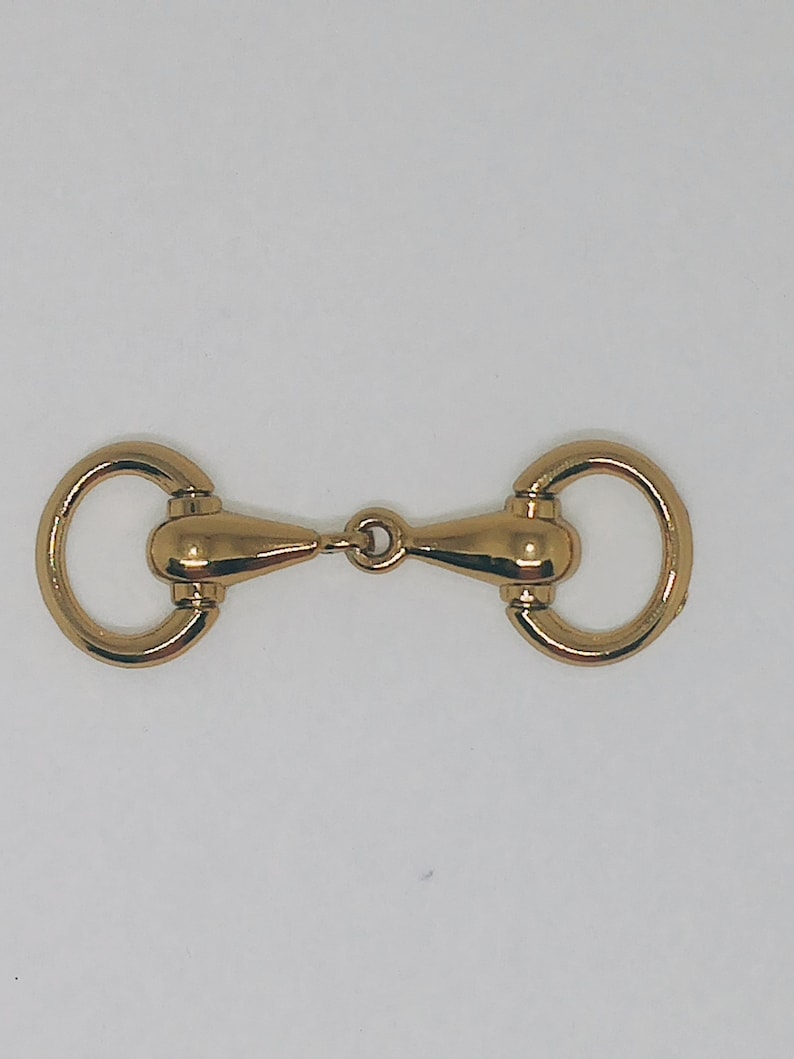 May include: Gold-toned metal horse bit buckle. The buckle has two rings and a centre piece with a rounded shape.
